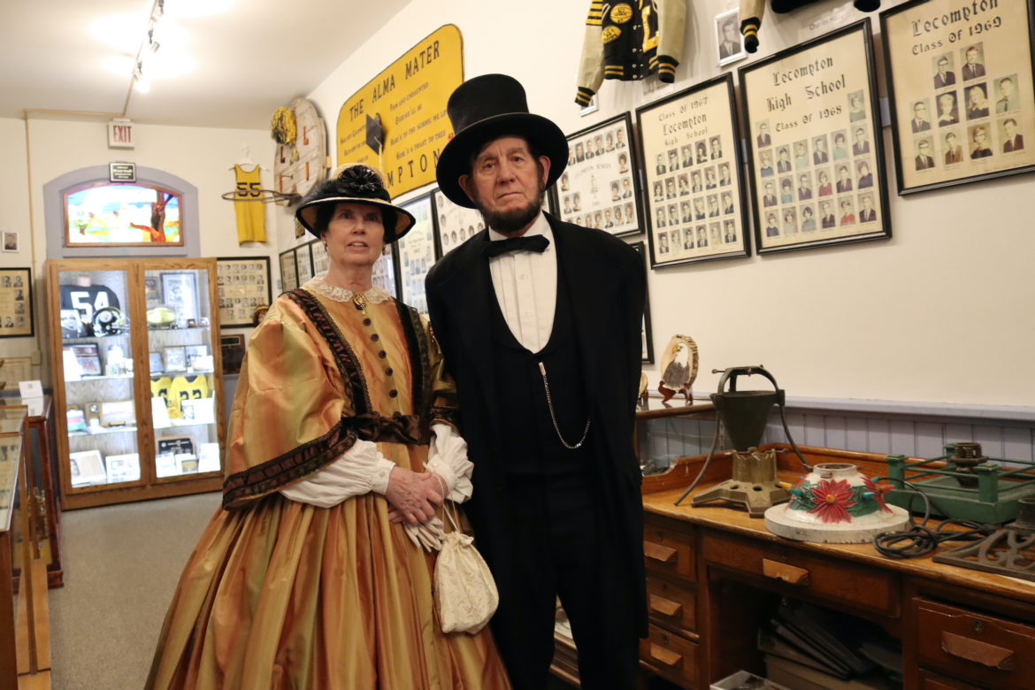 一名身穿19世纪金色连衣裙、戴着帽子和手套的女子站在一名身穿黑色西装、戴着礼帽的男子旁边。